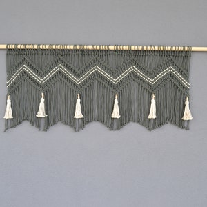 Könnte beinhalten: Ein grauer Makramee-Wandbehang mit Chevron-Muster und weißen Quasten. Der Behang ist an einer Holzstange mit Messing-Endkappen befestigt.