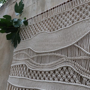 K&ouml;nnte beinhalten: Ein gro&szlig;er, cremefarbener Makramee-Wandbehang mit einem gewebten Muster aus Wellen und geometrischen Formen. Der Behang hat einen Fransenrand und ist an einer Wand mit einer Holzleiste befestigt.