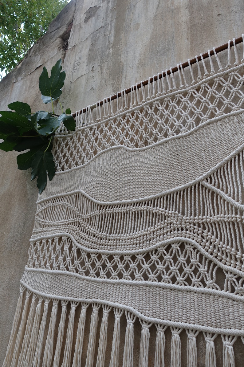 K&ouml;nnte beinhalten: Ein gro&szlig;er, cremefarbener Makramee-Wandbehang mit einem gewebten Muster aus Wellen und geometrischen Formen. Der Behang hat einen Fransenrand und ist an einer Wand mit einer Holzleiste befestigt.