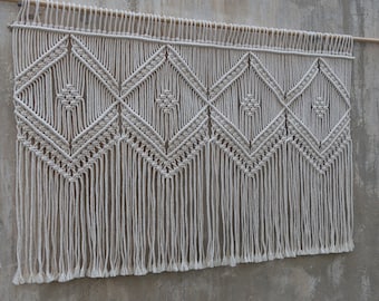 May include: A white macrame wall hanging with a geometric diamond pattern. The hanging is made of cotton cord and has a fringe at the bottom.