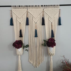 Puede incluir: Colgante de pared de macramé color crema con tres secciones, cada una con un diseño tejido, cuentas de madera y borlas azul marino. Debajo, cuelgan maceteros de macramé blanco con flores color burdeos.