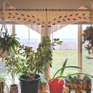 May include: A cream-coloured macrame window valance featuring a repeating leaf pattern with dark brown accents and a fringed bottom edge. The valance is hung on a black rod and spans across a wide window, positioned above several potted plants.