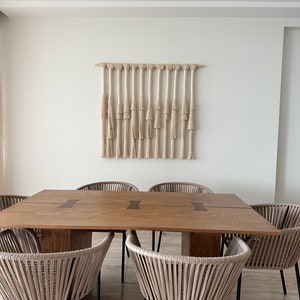 K&ouml;nnte beinhalten: Ein Esstisch aus Holz mit einer nat&uuml;rlichen Holzoptik und einem gewebten Makramee-Wandbehang dar&uuml;ber. Der Tisch hat eine rechteckige Form und ist von sechs gewebten St&uuml;hlen mit schwarzen Metallbeinen umgeben.