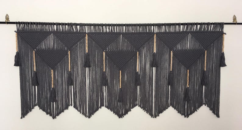May include: Gray macrame wall hanging with wooden beads and tassels. The hanging is made of a thick, textured cord and has a geometric pattern.
