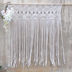 Puede incluir: Un tapiz de macramé blanco roto con un diseño geométrico y largas franjas. Cuelga de una barra de madera y está adornado con flores blancas y rosas en el lado izquierdo. La pared detrás es de color gris neutro.