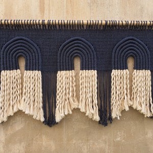 May include: A wall hanging with three black and white macrame arches. The arches are made with thick rope and have a fringe at the bottom.