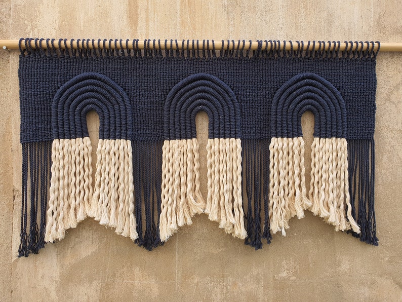 May include: A wall hanging with three black and white macrame arches. The arches are made with thick rope and have a fringe at the bottom.