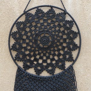Puede incluir: Atrapasueños de crochet negro con un diseño circular de macramé y una larga franja.