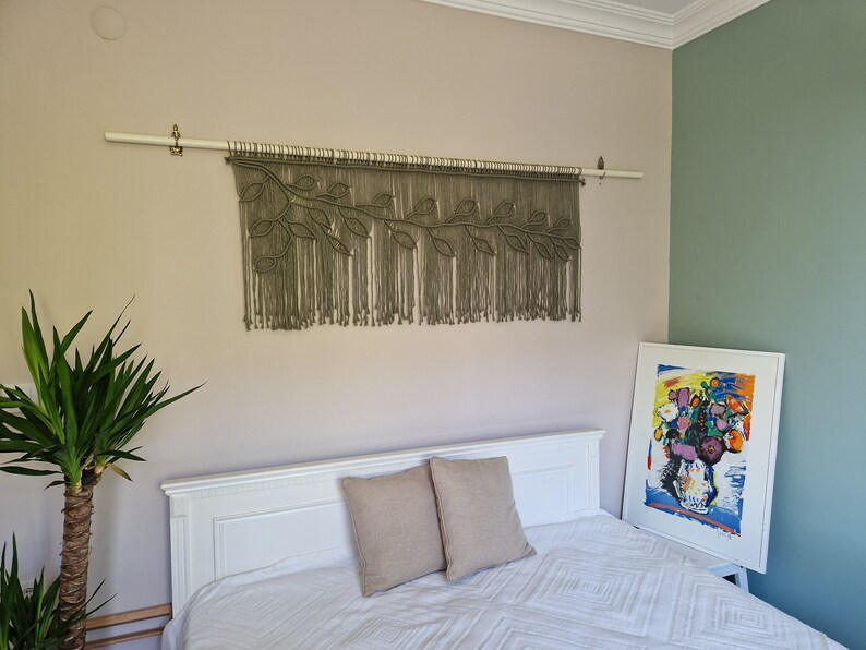 May include: A white bed with a white and beige patterned duvet and two beige throw pillows. A macrame wall hanging with a leafy design hangs above the bed. A framed abstract painting with colorful flowers is on the wall to the right of the bed.