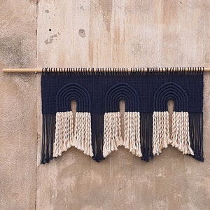 May include: A wall hanging with three navy blue and cream colored arches, with fringe on the bottom. The arches are made of woven rope and are hung on a wooden dowel.