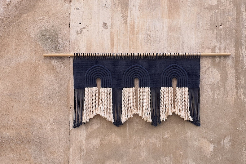 May include: A wall hanging with three navy blue and cream colored arches, with fringe on the bottom. The arches are made of woven rope and are hung on a wooden dowel.