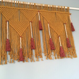 Può includere: Applique da parete in macramè giallo senape con motivi geometrici e frange. Presenta nappe bordeaux e fili di perline di legno. L'arredamento murale è montato su un'asta bianca contro un muro azzurro.