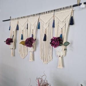 May include: Cream-coloured macrame wall hanging with woven design, featuring hanging planters with purple flowers. Navy blue tassels and wooden beads provide accents. The piece is mounted on a black metal rod.