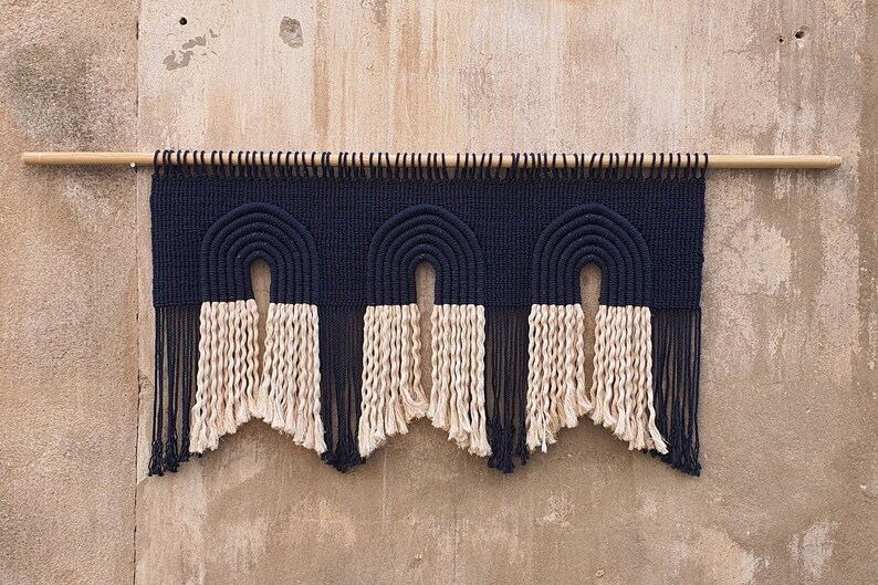 May include: A black and white macrame wall hanging with three arches. The arches are made of thick black cord and the bottom of the hanging has a fringe of white cord.