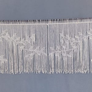 May include: A wide off-white macrame wall hanging featuring a horizontal design of interconnected leafy vine patterns. The piece is suspended from a thin black rod and consists of numerous vertical cord strands that create a fringe effect.