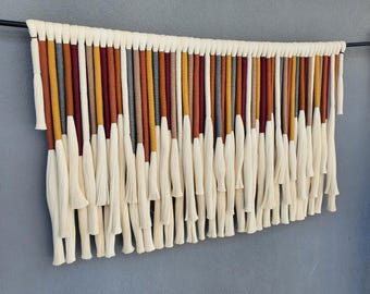 May include: A contemporary wall hanging with a striking design. It showcases vertical strands in cream, mustard yellow, burgundy, and grey. The strands are suspended from a black rod, creating a textured and visually appealing artwork.