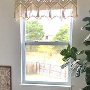 K&ouml;nnte beinhalten: Ein wei&szlig;er Makramee-Fenster&uuml;berhang mit Quasten h&auml;ngt &uuml;ber einem Fenster mit Blick auf einen Garten und einen Holzzaun.