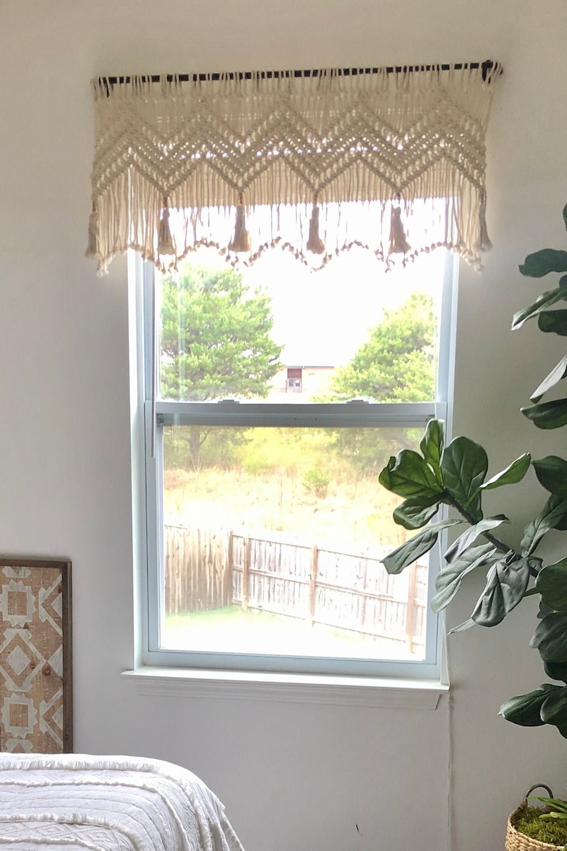 K&ouml;nnte beinhalten: Ein wei&szlig;er Makramee-Fenster&uuml;berhang mit Quasten h&auml;ngt &uuml;ber einem Fenster mit Blick auf einen Garten und einen Holzzaun.