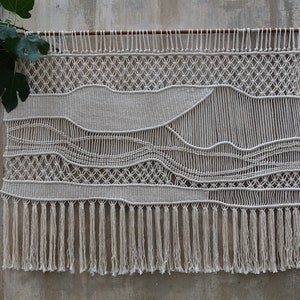 K&ouml;nnte beinhalten: Ein wei&szlig;er Makramee-Wandbehang mit geometrischem Muster und Fransen. Der Wandbehang ist aus nat&uuml;rlicher Baumwollschnur gefertigt und hat einen rustikalen, bohemian Stil.