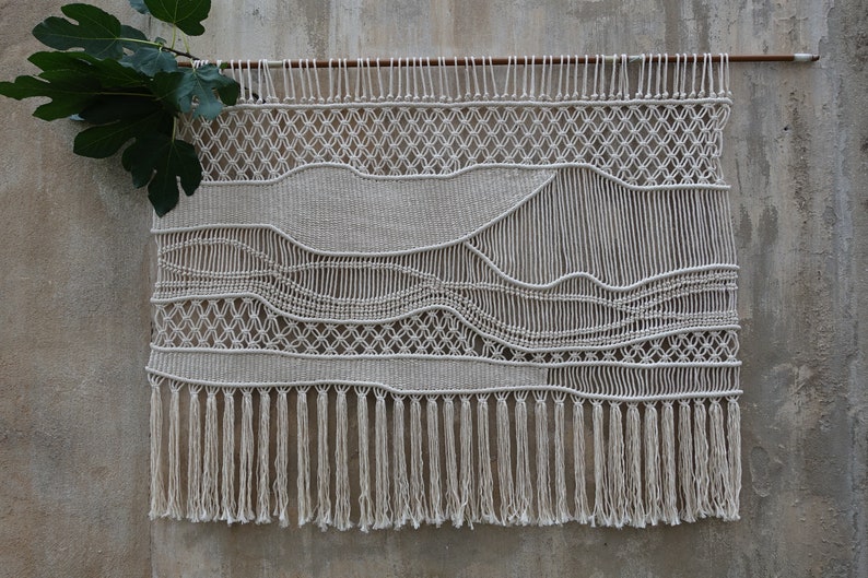 K&ouml;nnte beinhalten: Ein wei&szlig;er Makramee-Wandbehang mit geometrischem Muster und Fransen. Der Wandbehang ist aus nat&uuml;rlicher Baumwollschnur gefertigt und hat einen rustikalen, bohemian Stil.