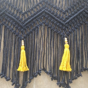 May include: A black macrame wall hanging with a geometric pattern and two yellow tassels.