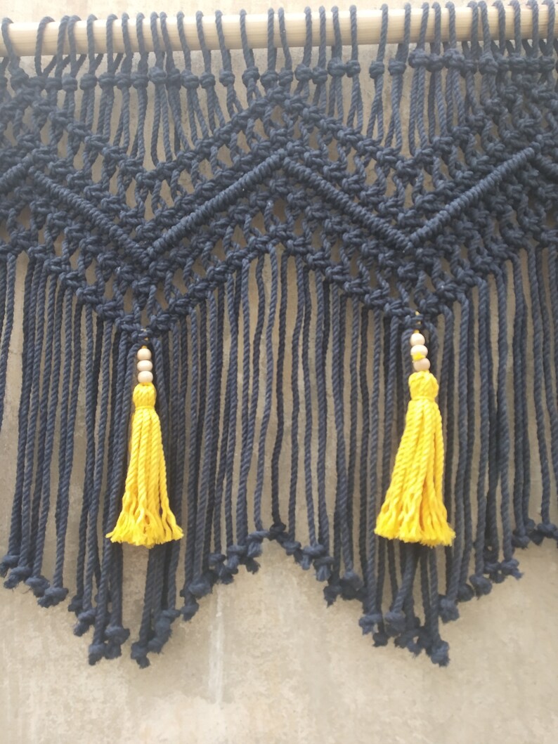 May include: A black macrame wall hanging with a geometric pattern and two yellow tassels.