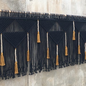 May include: Black and gold macrame wall hanging with geometric patterns and tassels. The hanging features white beads and is made with natural fibres.