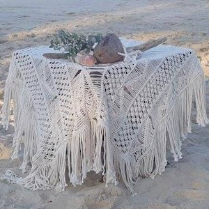 May include: A white crocheted table runner with fringe, featuring a delicate openwork pattern. The runner is draped over a wooden table and decorated with natural elements, including a coconut, seashells, and greenery.