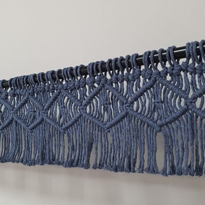 May include: A blue macrame wall hanging with a geometric diamond pattern and fringe. The handmade textile is suspended from a black rod, adding a touch of bohemian decor to any space. The color is a muted blue.