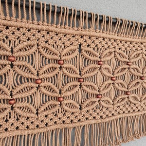 Op de afbeelding: Een beige macramé wandkleed met een geometrisch patroon en houten kralen. Het handgemaakte stuk heeft een reeks geknoopte ontwerpen en een franje aan de onderkant. De wanddecoratie is gemonteerd op een donkere metalen staaf.