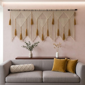 Könnte beinhalten: Ein großer cremefarbener Makramee-Wandbehang mit geometrischen Dreiecksmustern, Holzperlen und senfgelben Quasten, der an einer dunklen Stange hängt, kombiniert mit einem grauen Sofa, einem zylindrischen Makramee-Kissen und zwei Samtkissen.