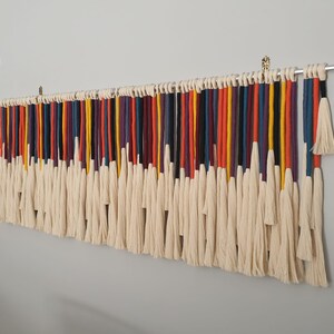 Puede incluir: Un tapiz de macram&eacute; de pared colorido con una variedad de colores que incluyen rojo, naranja, amarillo, azul, verde y morado. El tapiz est&aacute; hecho de hilo y tiene una franja en la parte inferior.