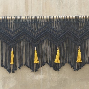May include: Black macrame wall hanging with a geometric pattern and yellow tassels. The hanging is attached to a wooden dowel.