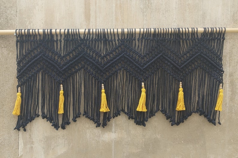May include: Black macrame wall hanging with a geometric pattern and yellow tassels. The hanging is attached to a wooden dowel.