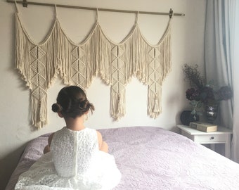 May include: A young girl sits on a bed with a white lace dress. Behind her is a macrame wall hanging with a cream color. The wall hanging is made of cotton cord and has a geometric pattern. The bed has a light purple comforter.