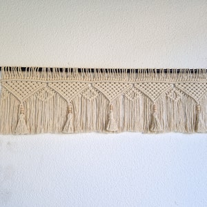 May include: A macrame wall hanging with a geometric pattern and tassels. The hanging is made of natural cotton cord and has a wooden dowel at the top.