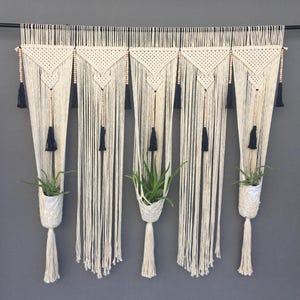 Puede incluir: Un tapiz de macramé con cinco paneles color crema, cada uno con un diseño triangular y flecos largos y verticales. Borlas azul oscuro y cuentas de madera acentúan el diseño. Debajo cuelgan maceteros de macramé blanco con plantas verdes.