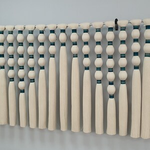 May include: A set of 20 white yarn tassels with teal and white striped bands and white beads. The tassels are hanging from a metal rod.