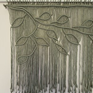 May include: A green macrame wall hanging with a leaf design. The hanging is made with thick cotton cord and has a fringe at the bottom.