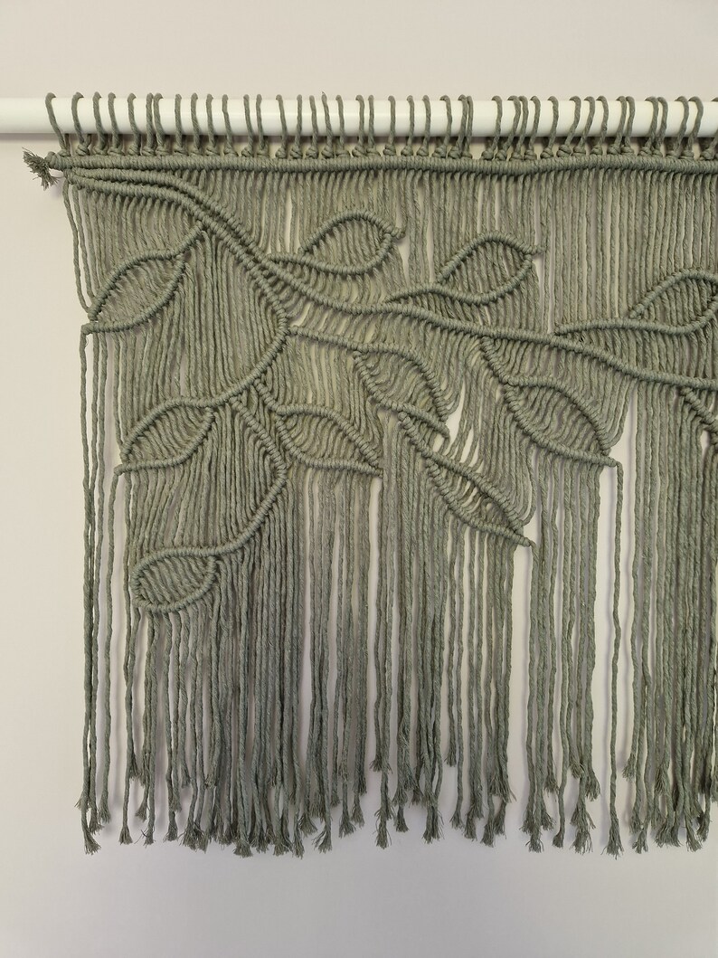 May include: A green macrame wall hanging with a leaf design. The hanging is made with thick cotton cord and has a fringe at the bottom.