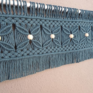 Pode incluir: Tapeçaria de parede em macramé azul-petróleo com um padrão floral geométrico e contas de madeira. O têxtil artesanal apresenta uma franja na parte inferior e é montado numa haste branca. Esta peça decorativa adiciona um toque boémio.