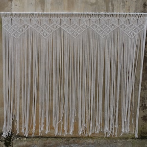 May include: White macrame wall hanging with a geometric pattern. The hanging is made of cotton cord and has a fringe at the bottom.