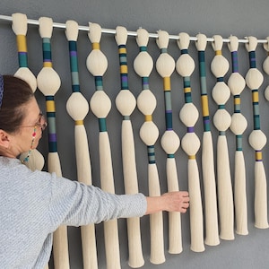 May include: A wall hanging with multiple vertical strands. Each strand features a cream-colored tassel, a central white orb, and sections of teal, yellow, and purple. The piece is mounted on a silver rod against a gray wall.