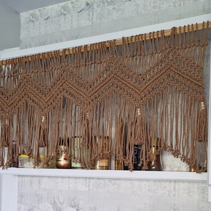 May include: A brown macrame wall hanging with a geometric pattern and wooden dowel rod. The hanging has small wooden beads and is hung above a shelf with jars and bottles.