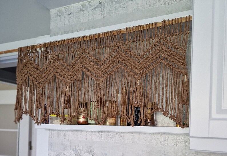 May include: A brown macrame wall hanging with a geometric pattern and wooden dowel rod. The hanging has small wooden beads and is hung above a shelf with jars and bottles.