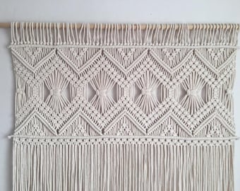 May include: A white macrame wall hanging with a geometric pattern and fringe. The hanging is made of cotton cord and is attached to a wooden dowel.