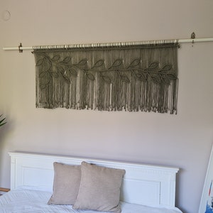 May include: A macrame wall hanging with a leafy design in a muted green color. The hanging is made of natural fibers and is attached to a wooden dowel rod.
