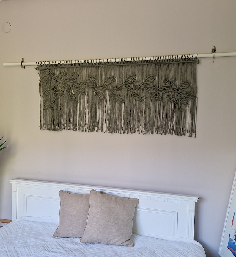 May include: A macrame wall hanging with a leafy design in a muted green color. The hanging is made of natural fibers and is attached to a wooden dowel rod.