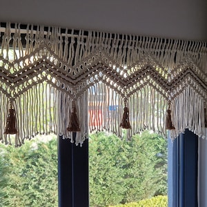 May include: A white macrame curtain valance with brown tassels hangs above a window with a view of green trees.