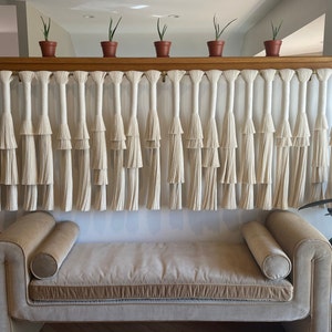 K&ouml;nnte beinhalten: Ein wei&szlig;er Makramee-Wandbehang mit mehreren Reihen von Quasten, der &uuml;ber einem hellbraunen Sofa mit zwei Kissen h&auml;ngt. Der Wandbehang ist an einem Holzregal mit kleinen Topfpflanzen befestigt.